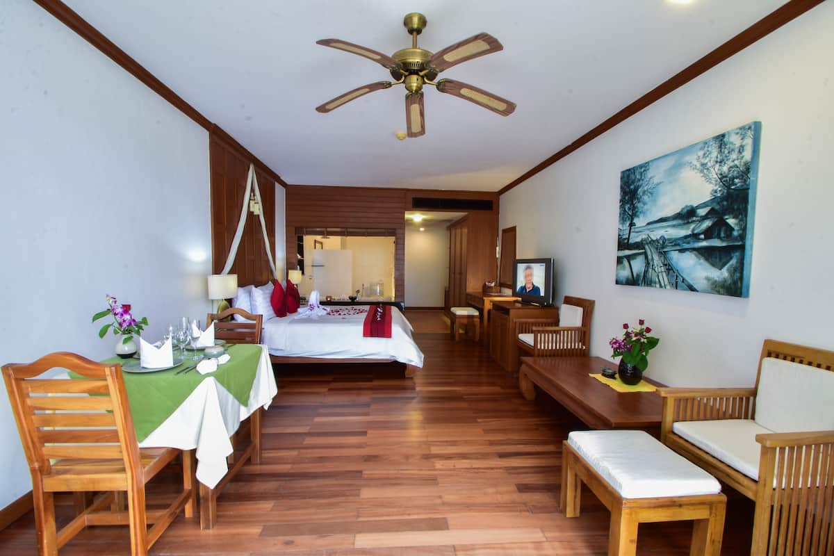 Une chambre d’hôtel avec un lit, des meubles en bois, une table à manger dressée pour deux personnes, un ventilateur de plafond et un tableau accroché au mur.