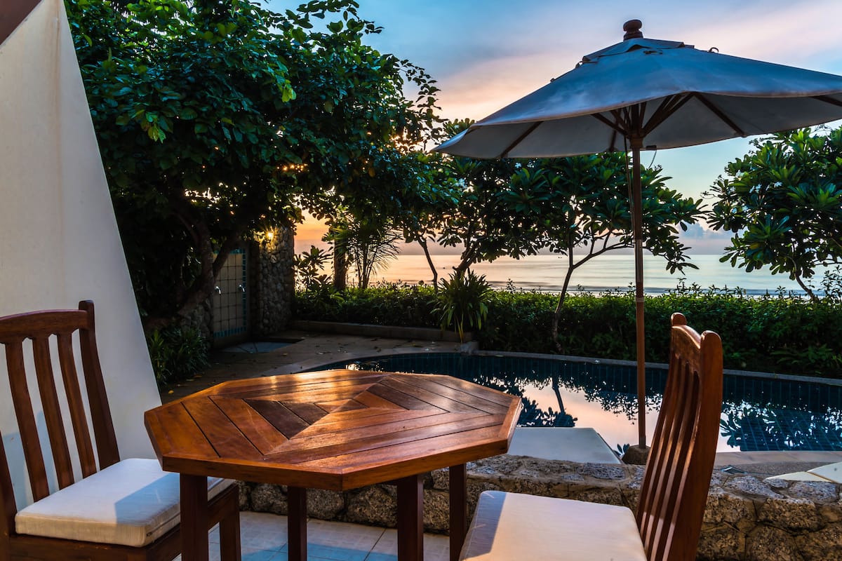 Une table et des chaises en bois, placées au bord d’une piscine, avec un parasol, entourées de verdure et offrant une vue sur la mer.