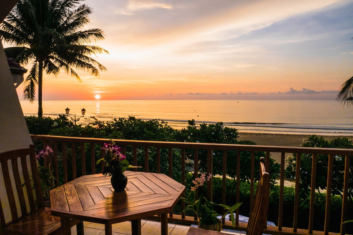 Une table en bois sur laquelle est posé un vase de fleurs, sur un balcon donnant sur un coucher de soleil sur l’océan.