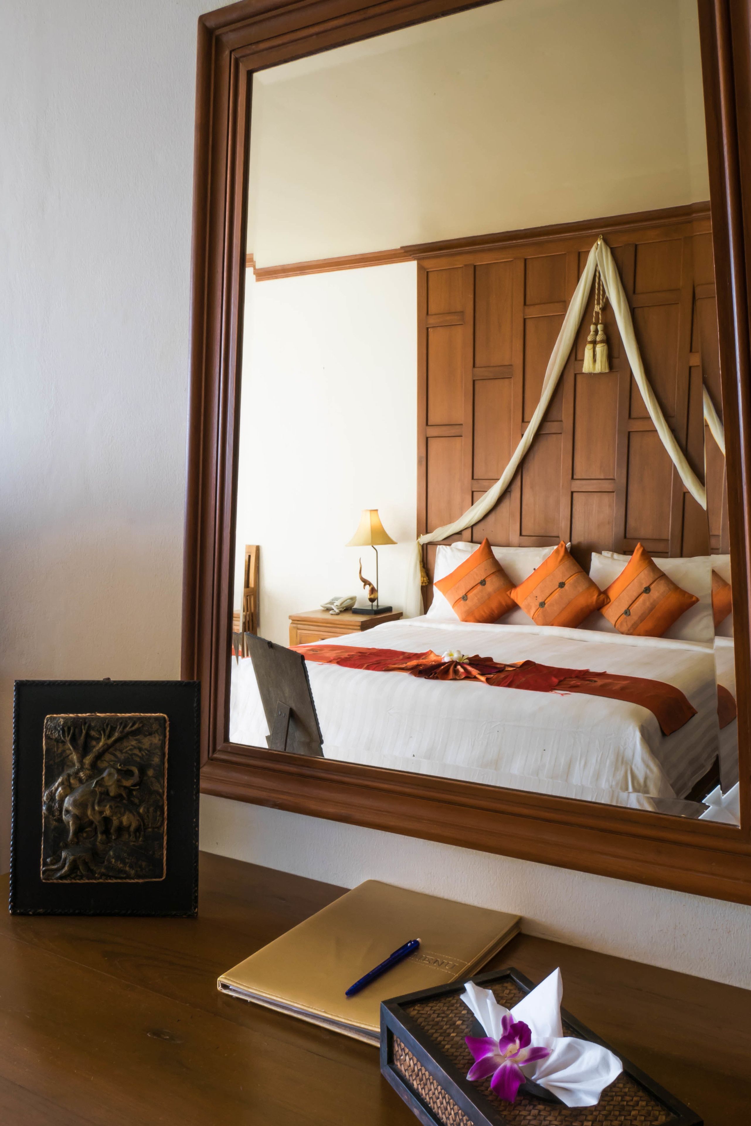 A hotel room with a large mirror reflecting a bed with orange pillows, a bedside table with a lamp, and a framed picture on the desk.