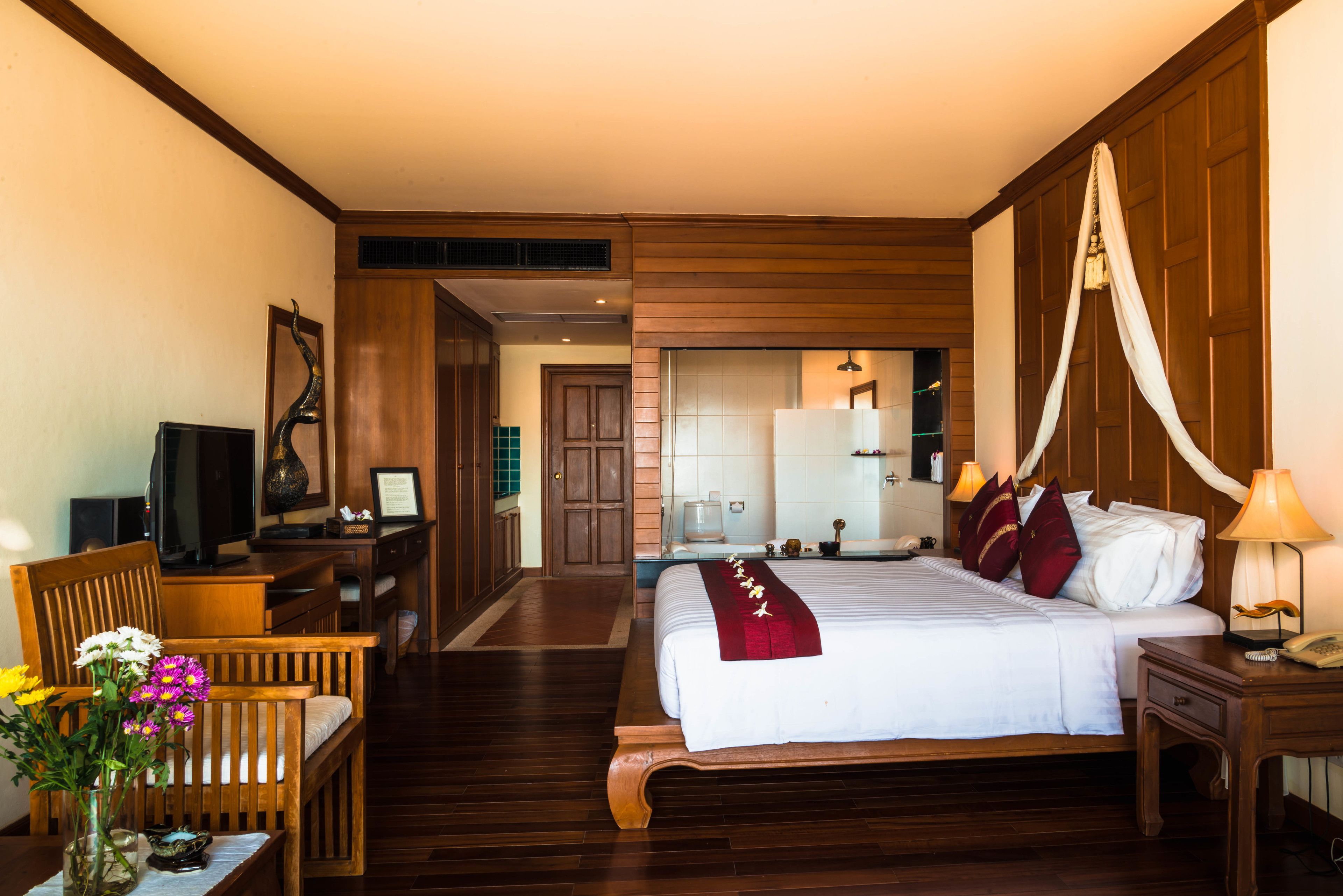 Una habitación de hotel con cama, mesita de noche, lámpara, silla, escritorio, televisor y un florero con flores.