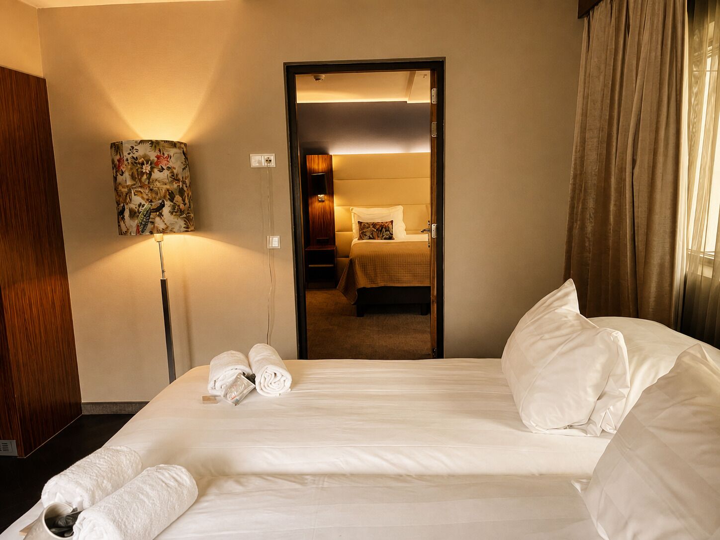Quarto de hotel com uma cama grande, abajur, espelho e vista para outro quarto.