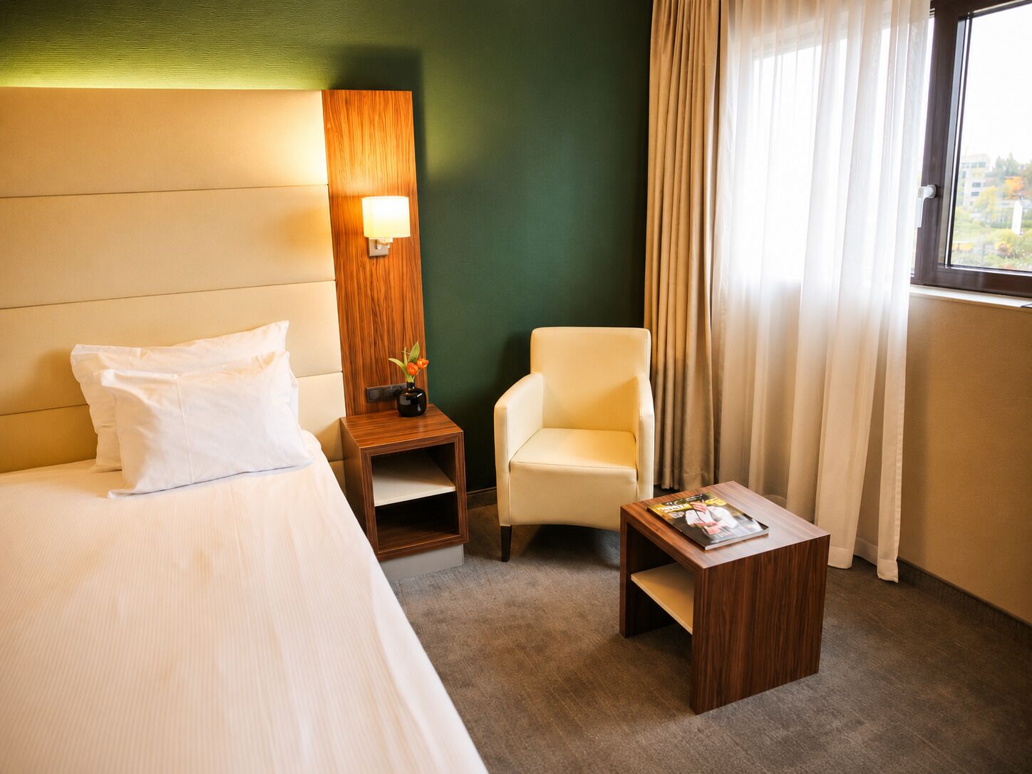 A hotel room with a bed, a chair, a small table, a nightstand, and a lamp.