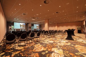 Un salón de conferencias con filas de sillas, una alfombra estampada con motivos florales y una pantalla de proyector.