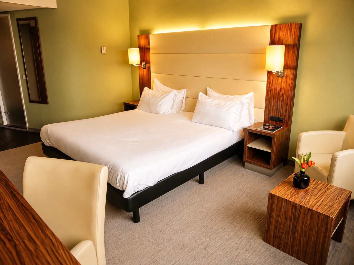 Una habitación de hotel con una cama grande, dos mesitas de noche, una silla y una pequeña mesa con un jarrón.