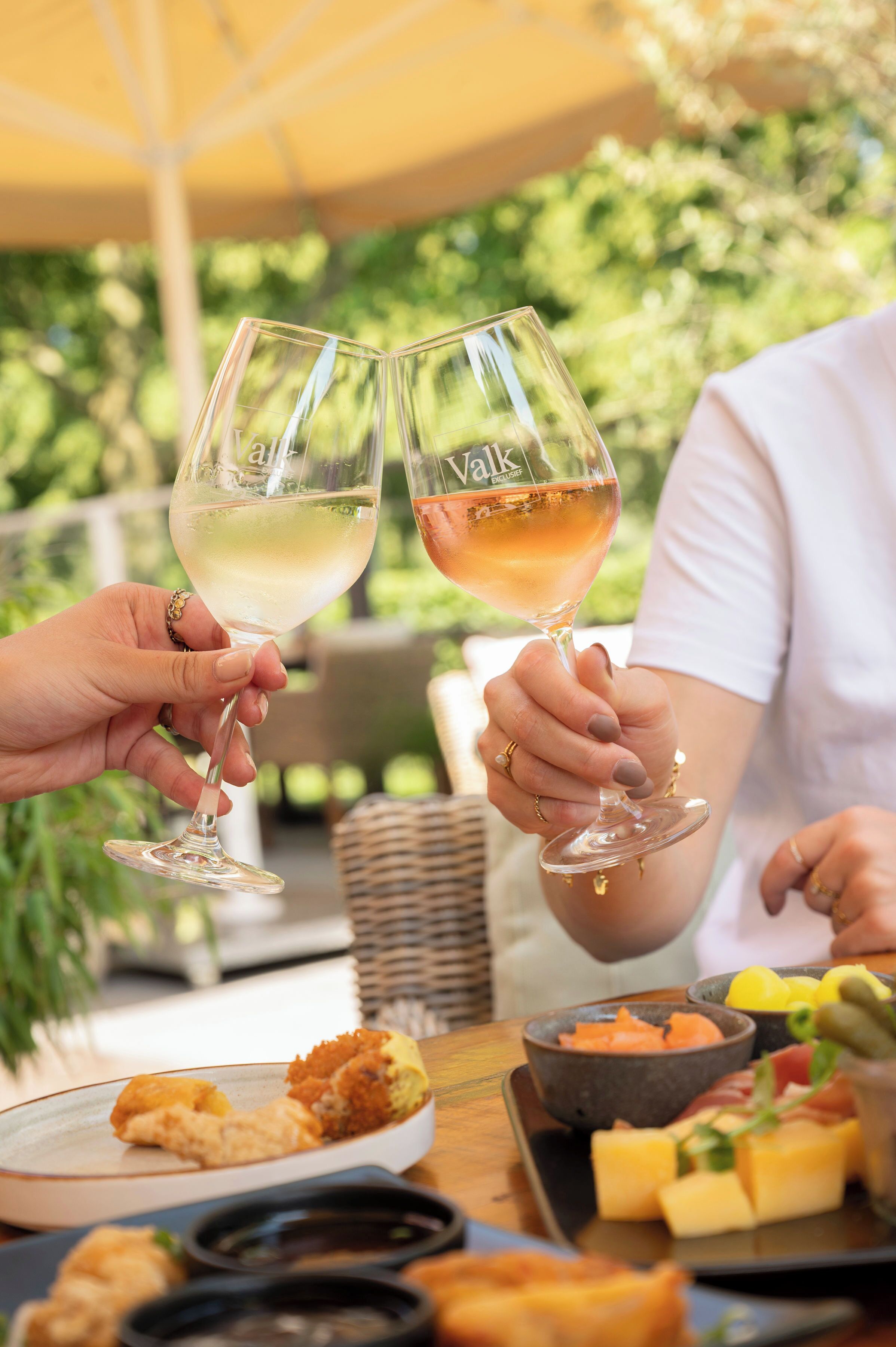 Two people toasting with wine glasses, a table with food and a wicker chair.