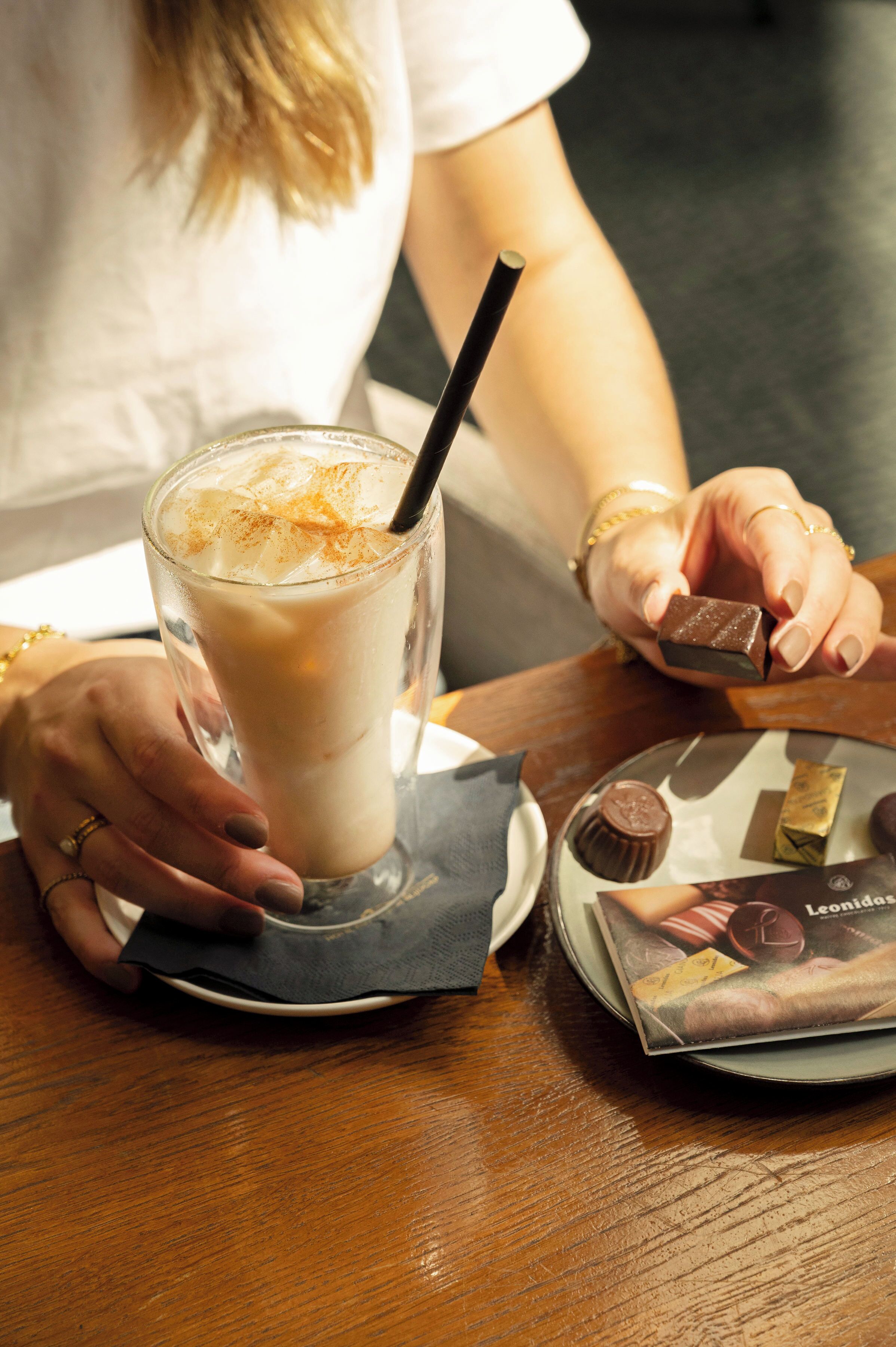 A person holding a glass of a beverage with a straw, a plate with chocolate pieces, and a chocolate bar on a table.