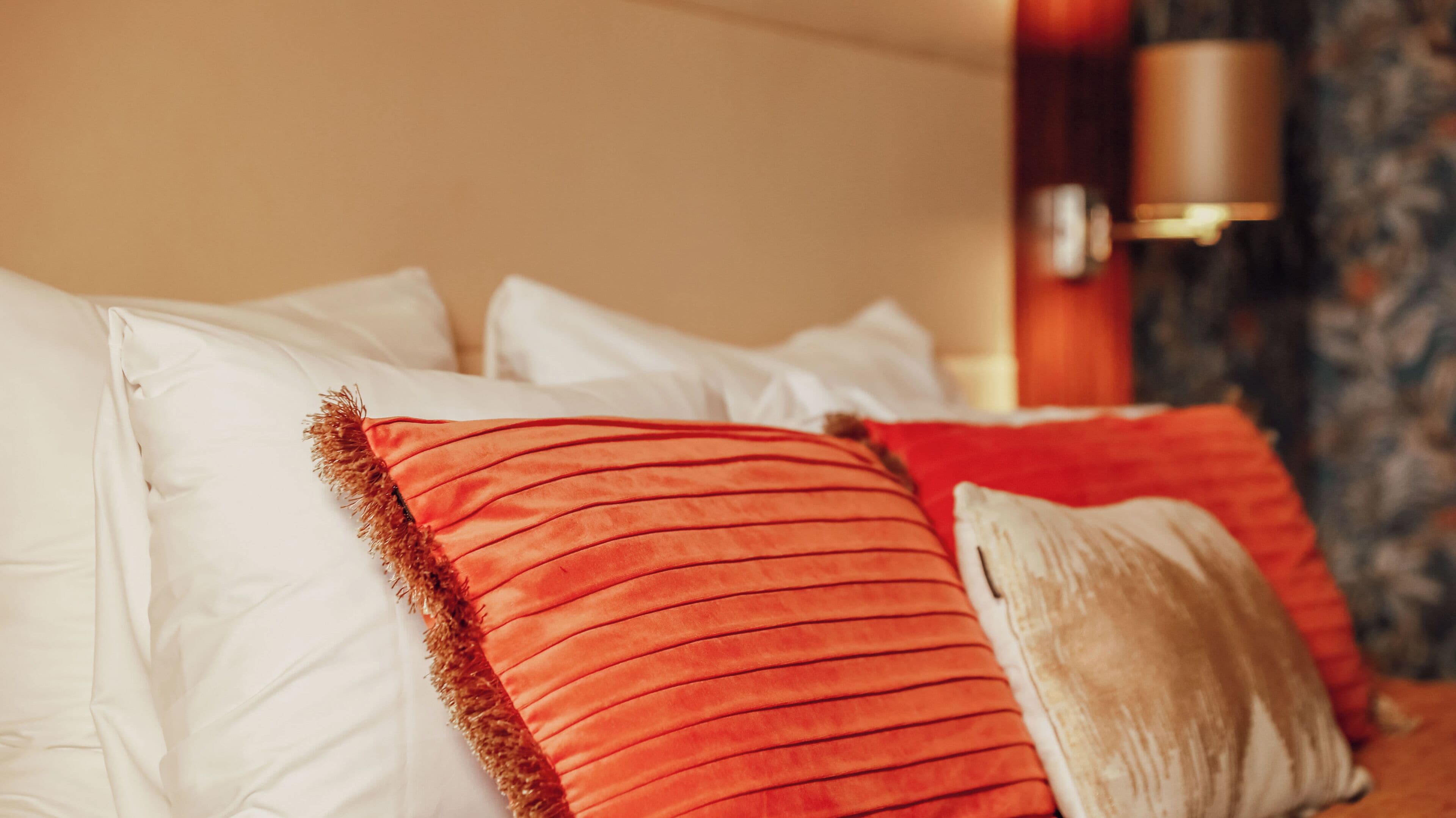 Una cama con almohadas blancas y rojas, una pared color beige y un papel tapiz estampado al fondo.