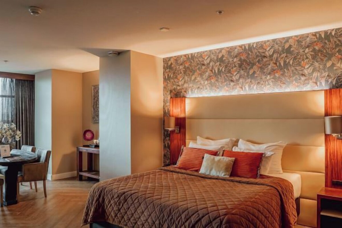 A hotel room with a large bed, a headboard with a floral wallpaper, and a bedside table with lamps.