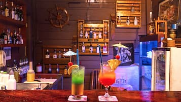 A bar area with two drinks on the counter, a ship's wheel on the wall, and shelves with bottles.