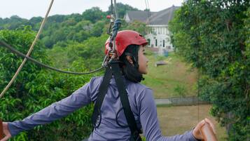 A person wearing a helmet and harness, holding onto a rope, with a house and greenery in the background.
