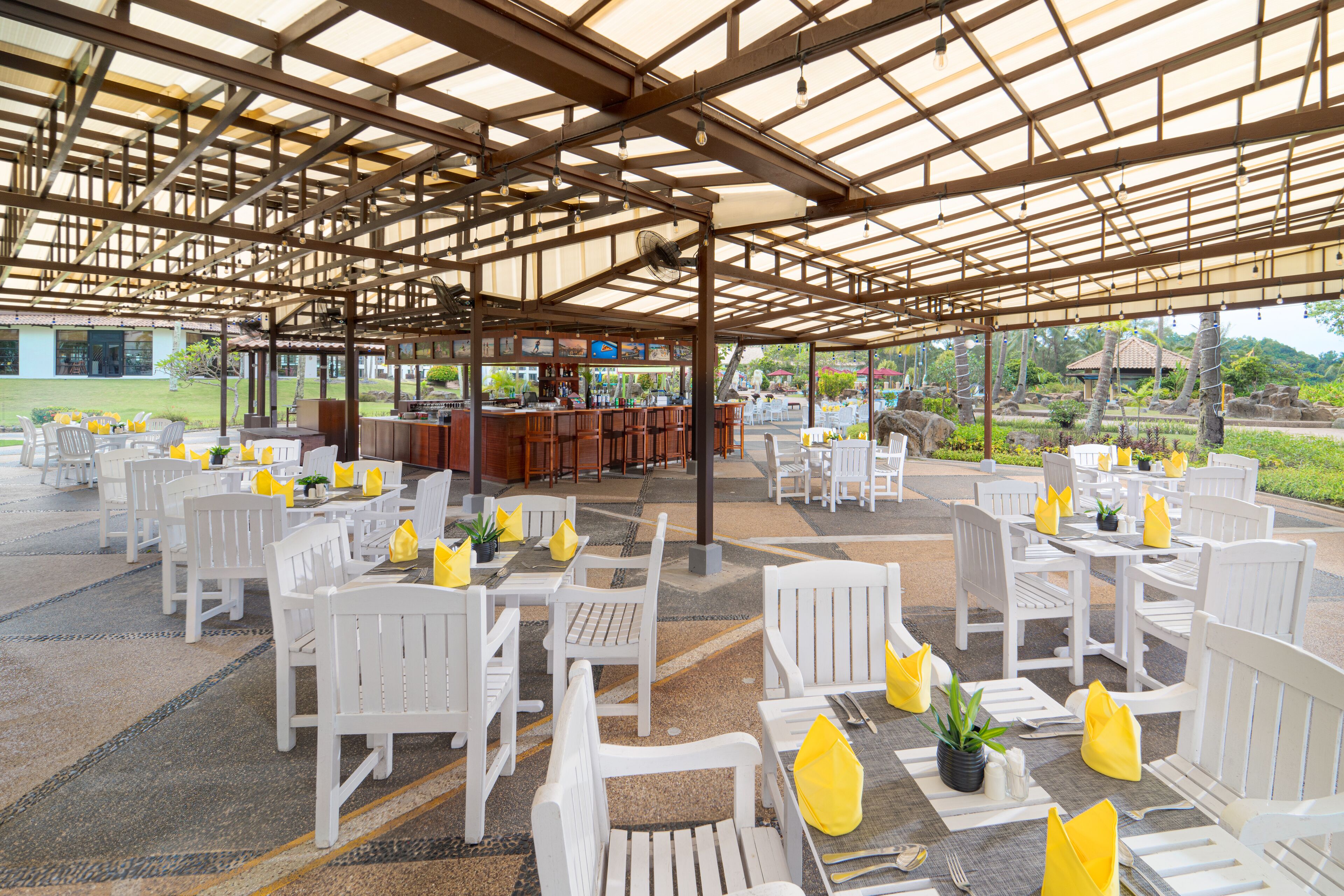 An outdoor covered seating area with white chairs and yellow cushions, a bar, and a view of the surrounding landscape.