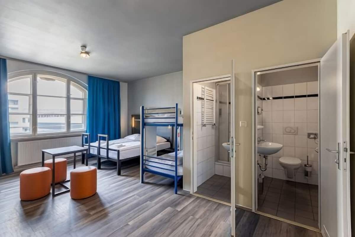 A room with bunk beds, a window with curtains, a table, and a bathroom with a sink and toilet.