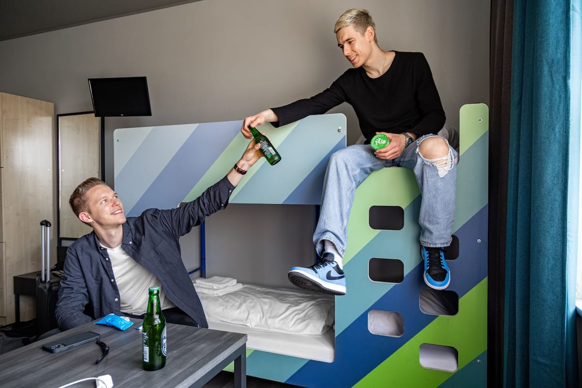 A bunk bed with a person sitting on the lower bunk and another standing on the upper bunk, holding a bottle.