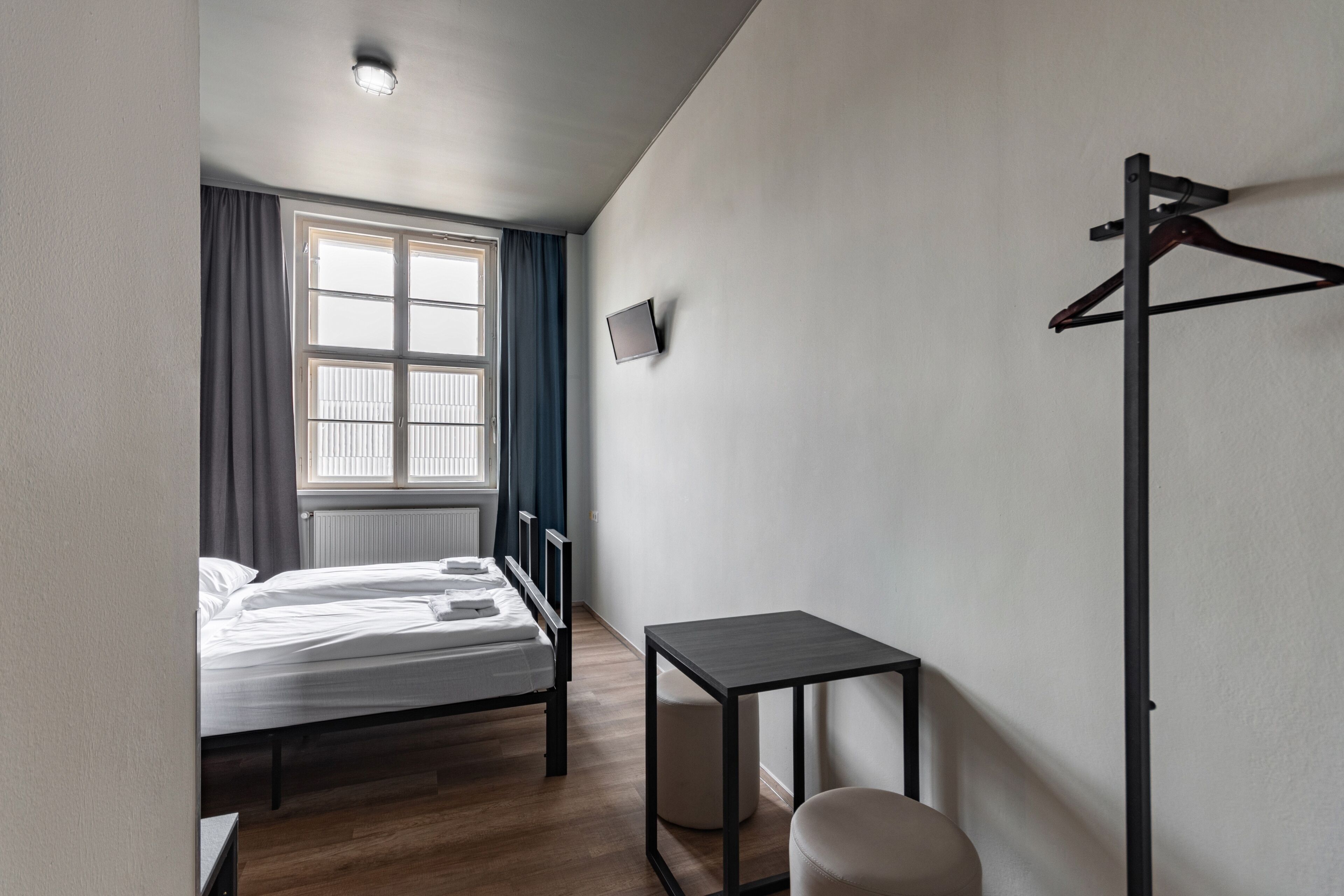 Ein Hotelzimmer mit Bett, kleinem Tisch, Bank, Kleiderständer und einem Fenster mit Vorhängen.