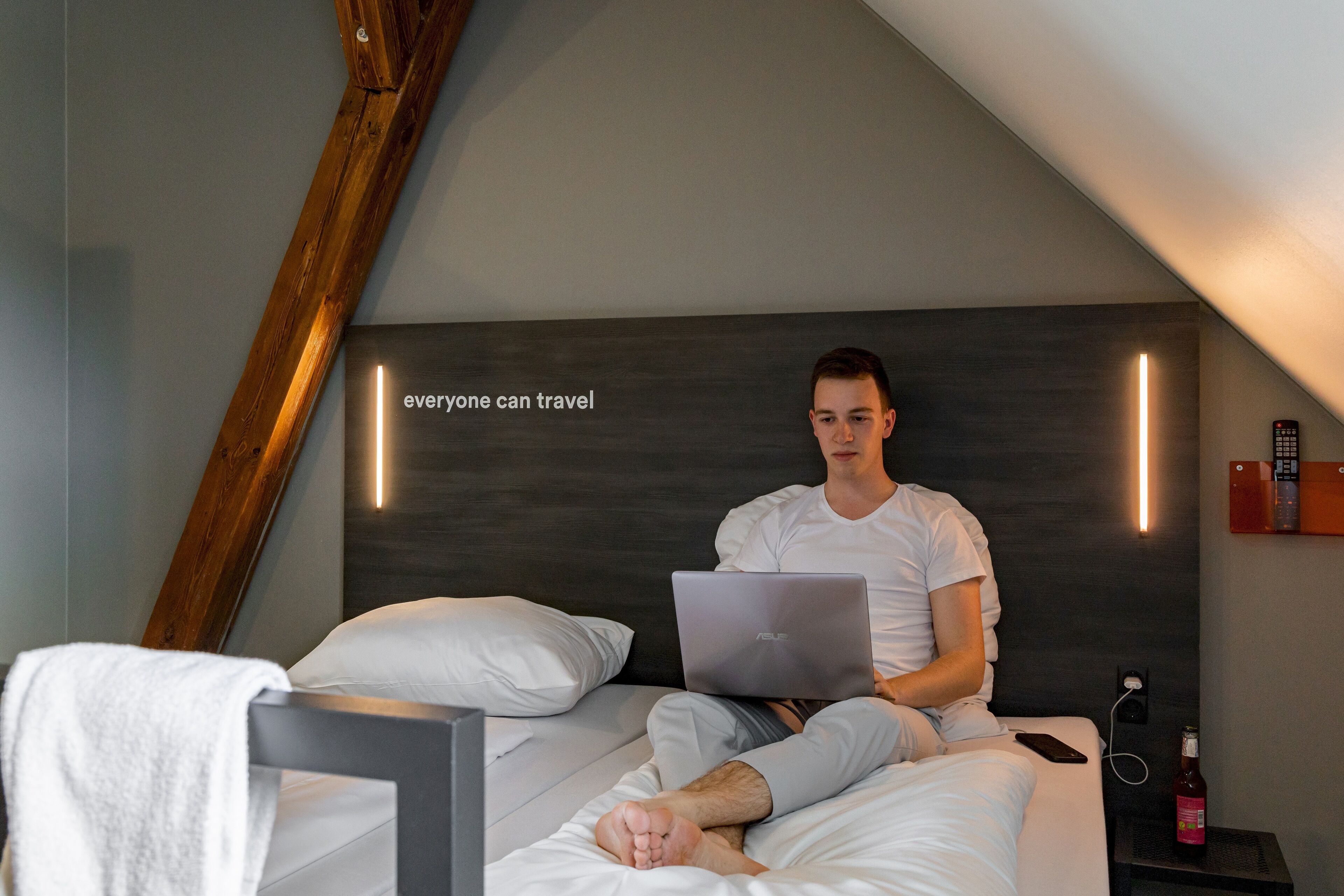 Une personne est assise sur un lit, avec un ordinateur portable, une télécommande et une bouteille sur une table de chevet.