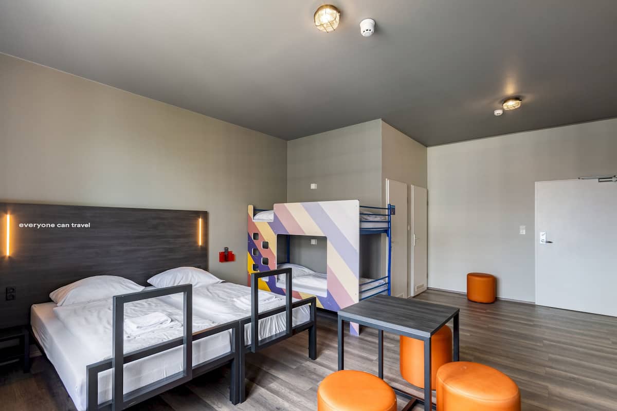 A modern hotel room with a large bed, a bunk bed, a small table, and orange ottomans.
