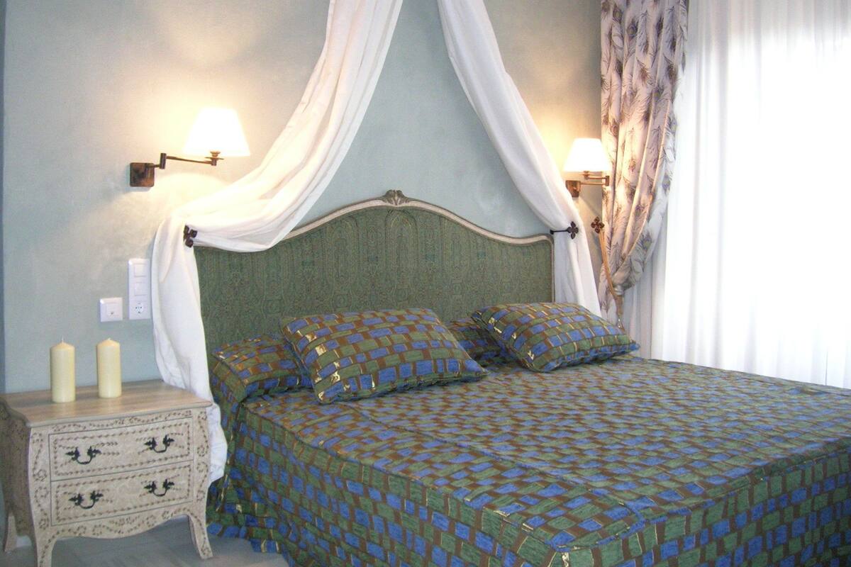 A bedroom with a four-poster bed, two bedside lamps, a nightstand with candles, and a window with curtains.