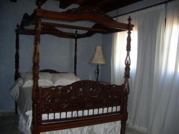 A four-poster bed with intricate carvings, a white bedspread, and a lamp on a bedside table in a room with white curtains.