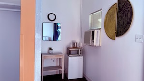 A small, compact room with a white wall, a wooden vanity, a microwave, a small refrigerator, a mirror, and a wall-mounted air conditioner.