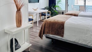 A modern hotel room with two beds, a desk, a TV, and a large window with a view of the beach.