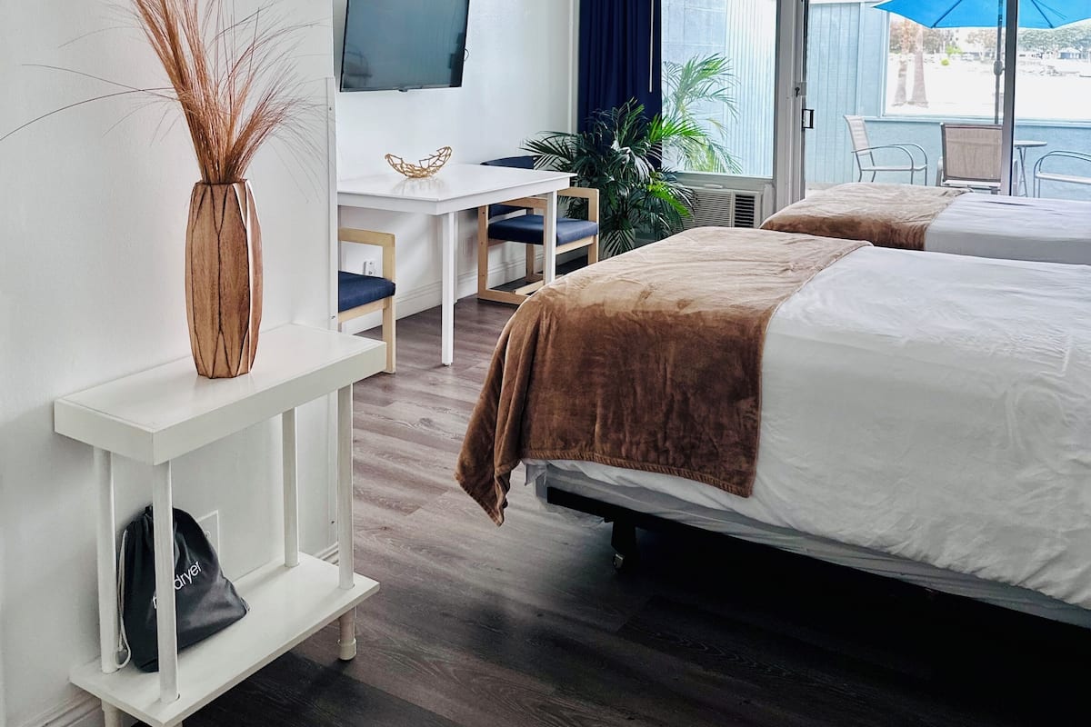 A modern hotel room with two beds, a desk, a TV, and a large window with a view of the beach.