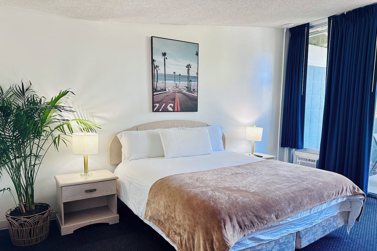 A hotel room with a bed, two bedside tables, a lamp, a potted plant, and a painting of a beach scene.