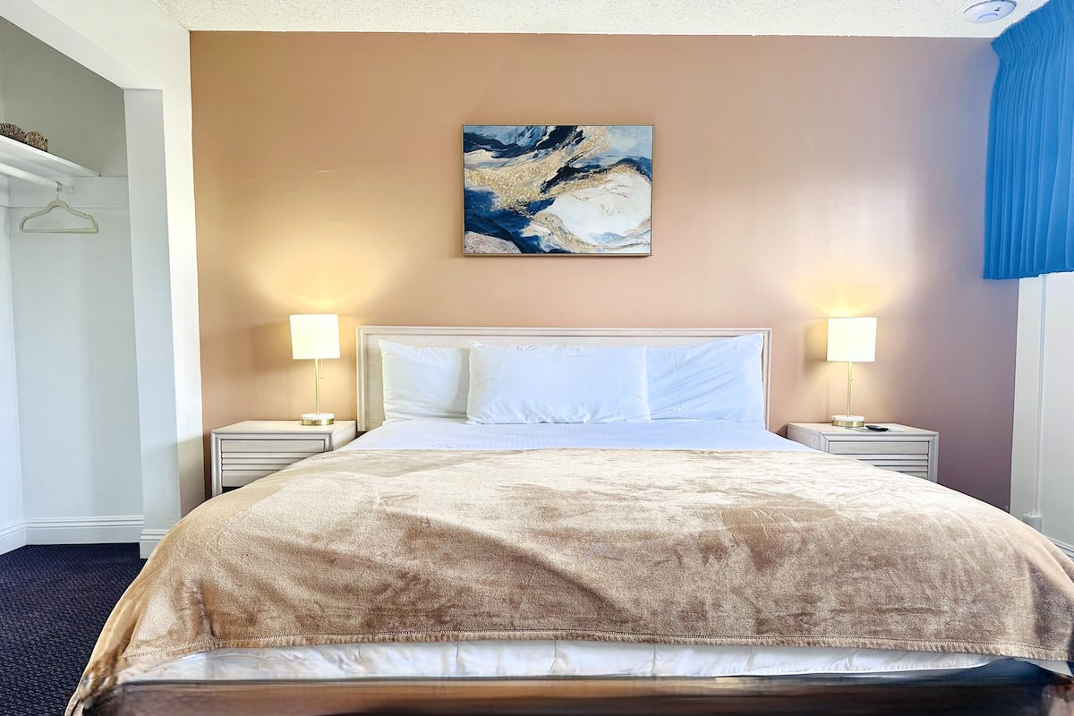 A hotel room with a large bed, two bedside tables with lamps, a painting on the wall, and a blue curtain.