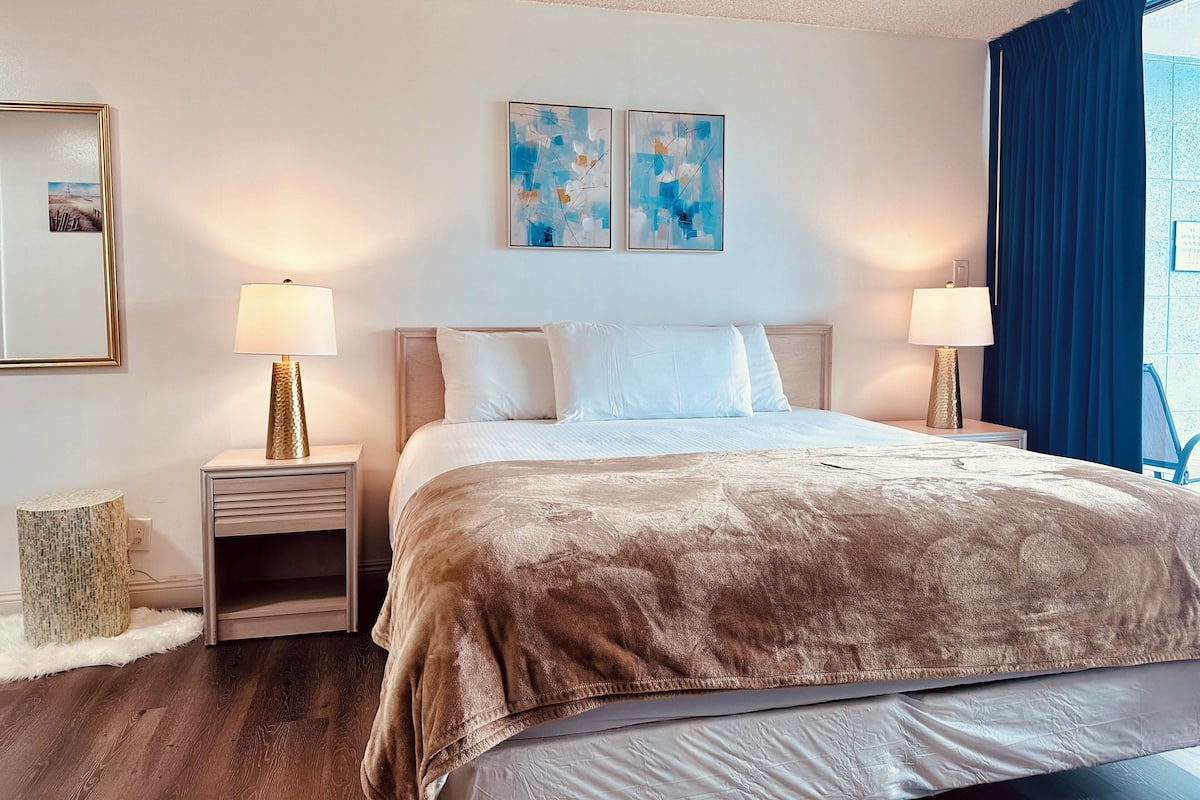 A hotel room with a large bed, two bedside lamps, a small side table, a decorative lamp, a framed picture, and abstract art on the wall.
