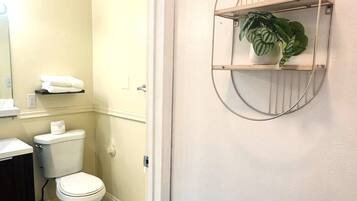 Un baño con inodoro, lavamanos y espejo. Hay una planta en un estante y un perchero para toallas.