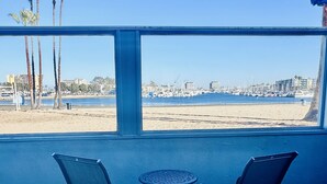 A patio with two chairs and a table, overlooking a beach and marina with boats and palm trees.