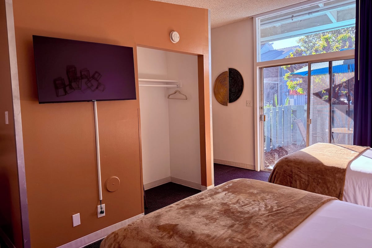 A hotel room with two beds, a TV mounted on the wall, a coat rack, and a sliding glass door leading to a balcony with a view of the outside.