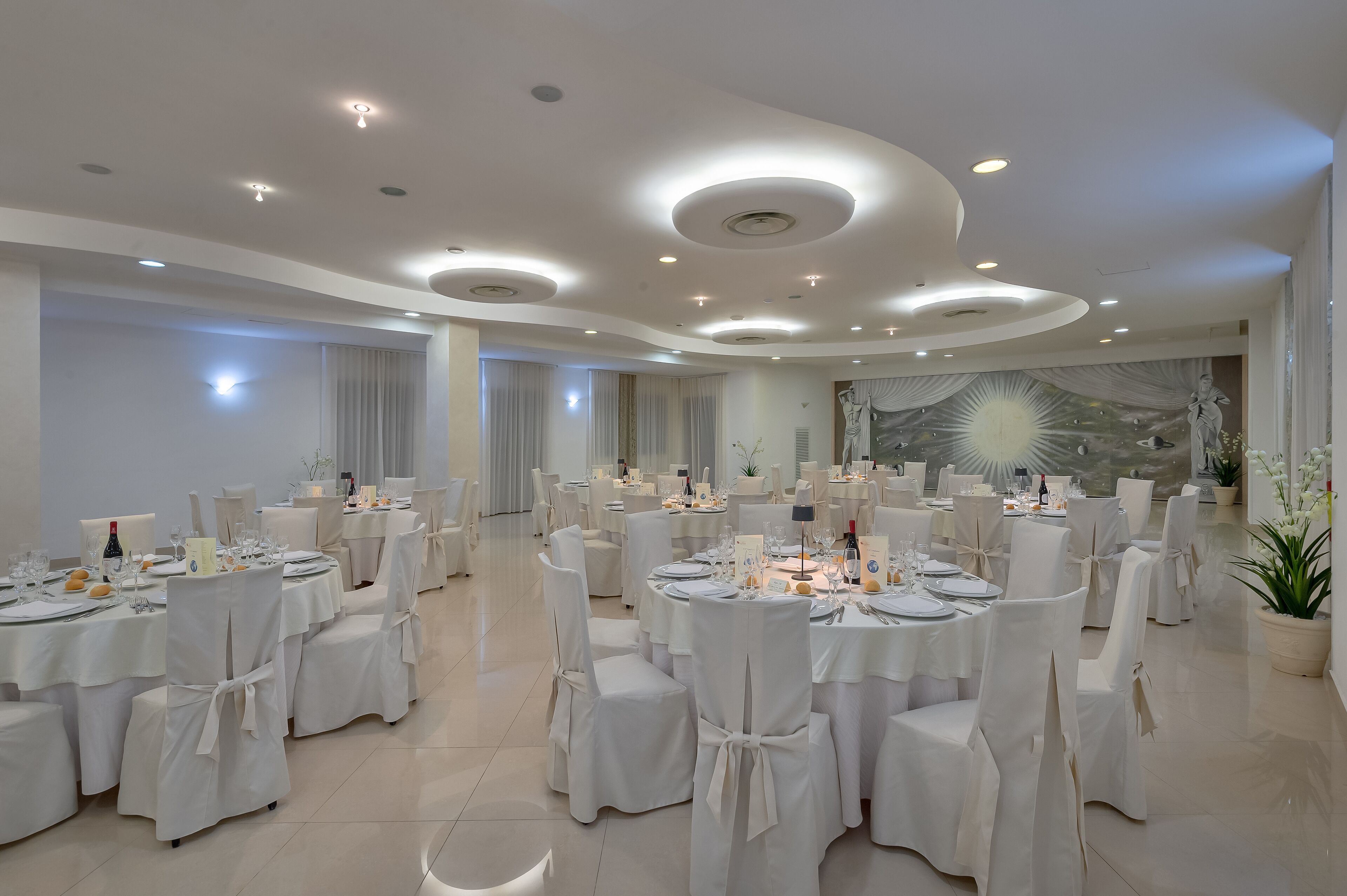 A banquet hall with round tables, white chairs, and a mural on the wall.