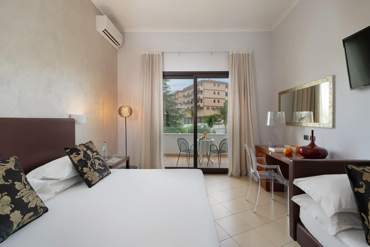 Une chambre d’hôtel avec un grand lit, un bureau avec une chaise, une télévision et un balcon offrant une vue sur les bâtiments et la verdure.