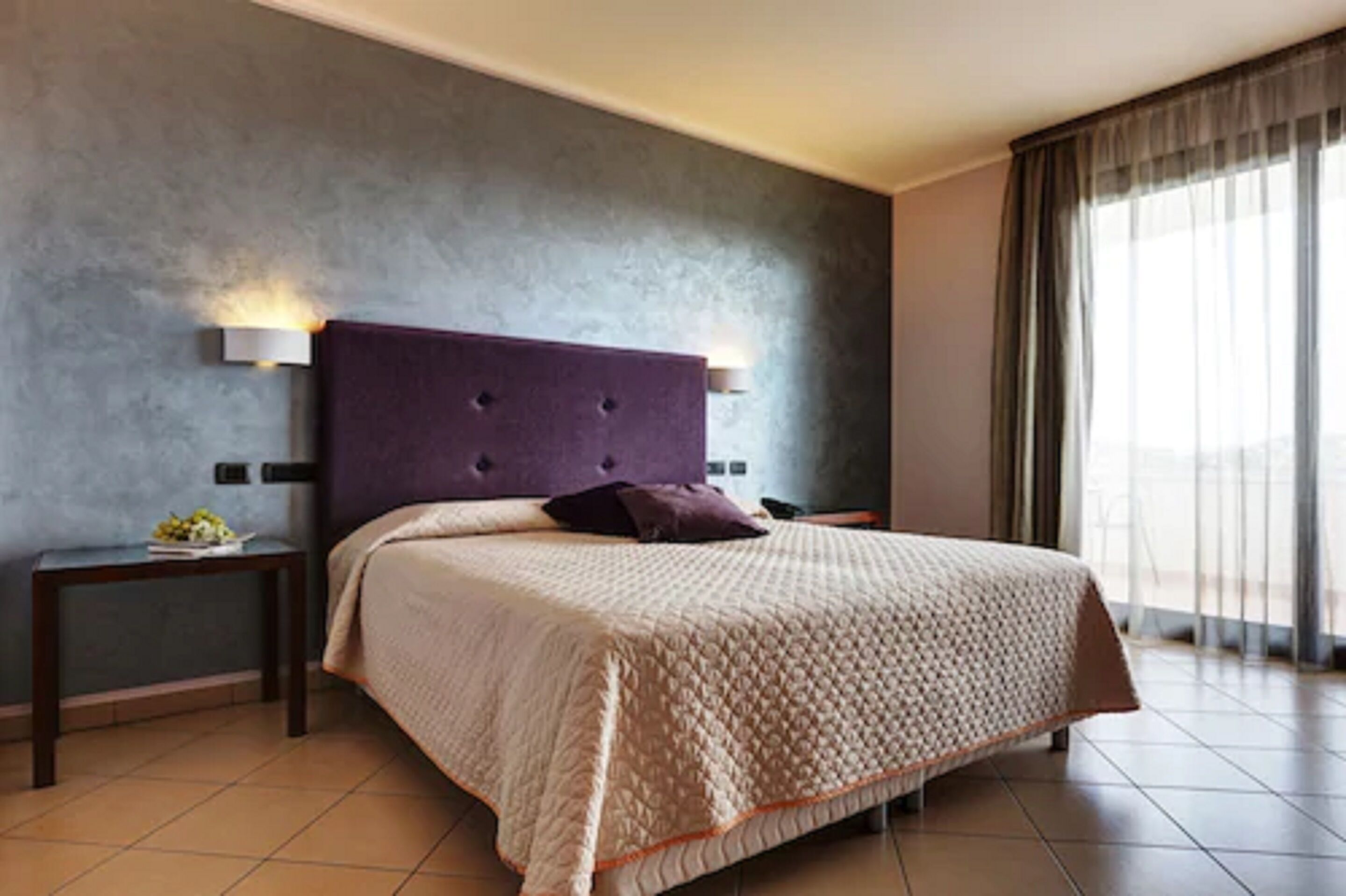 Ein Hotelzimmer mit einem großen Bett, einem Kopfteil mit Wandleuchten, einem Nachttisch mit Lampe und Blick ins Freie durch ein Fenster.