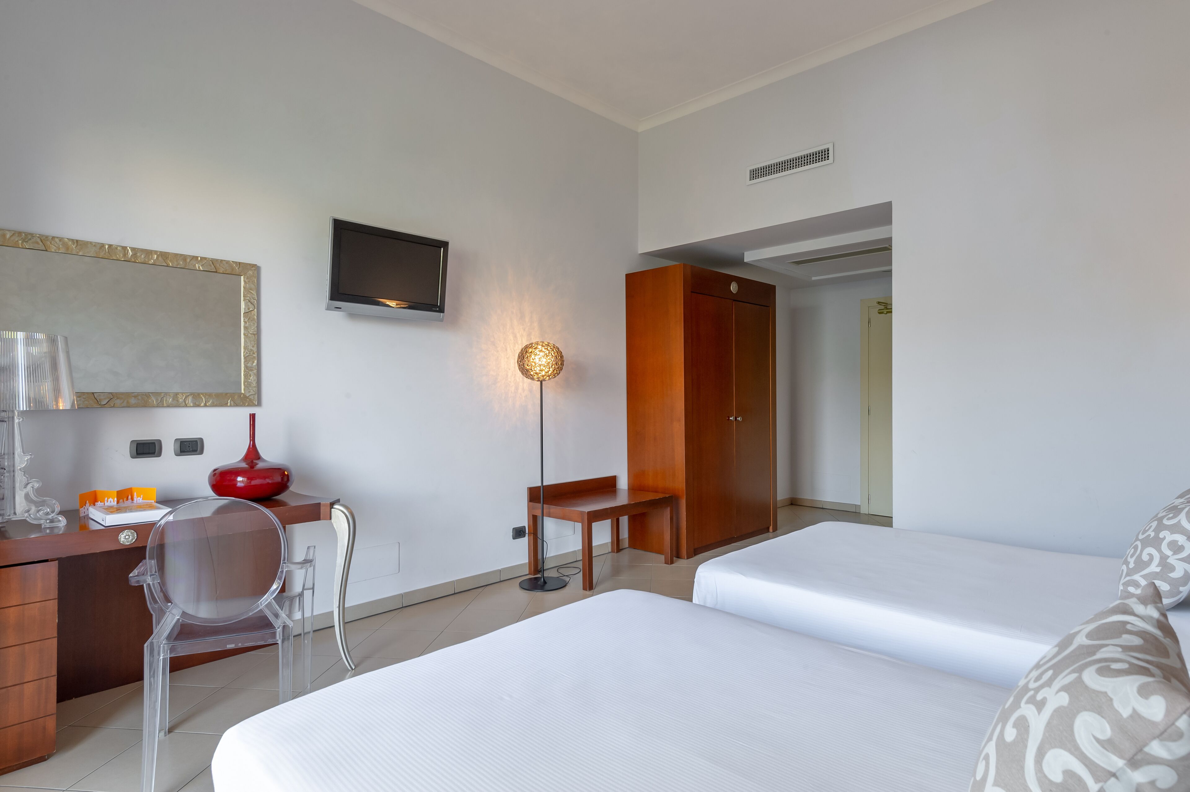 Quarto de hotel com cama, escrivaninha, cadeira, televisão e guarda-roupa.