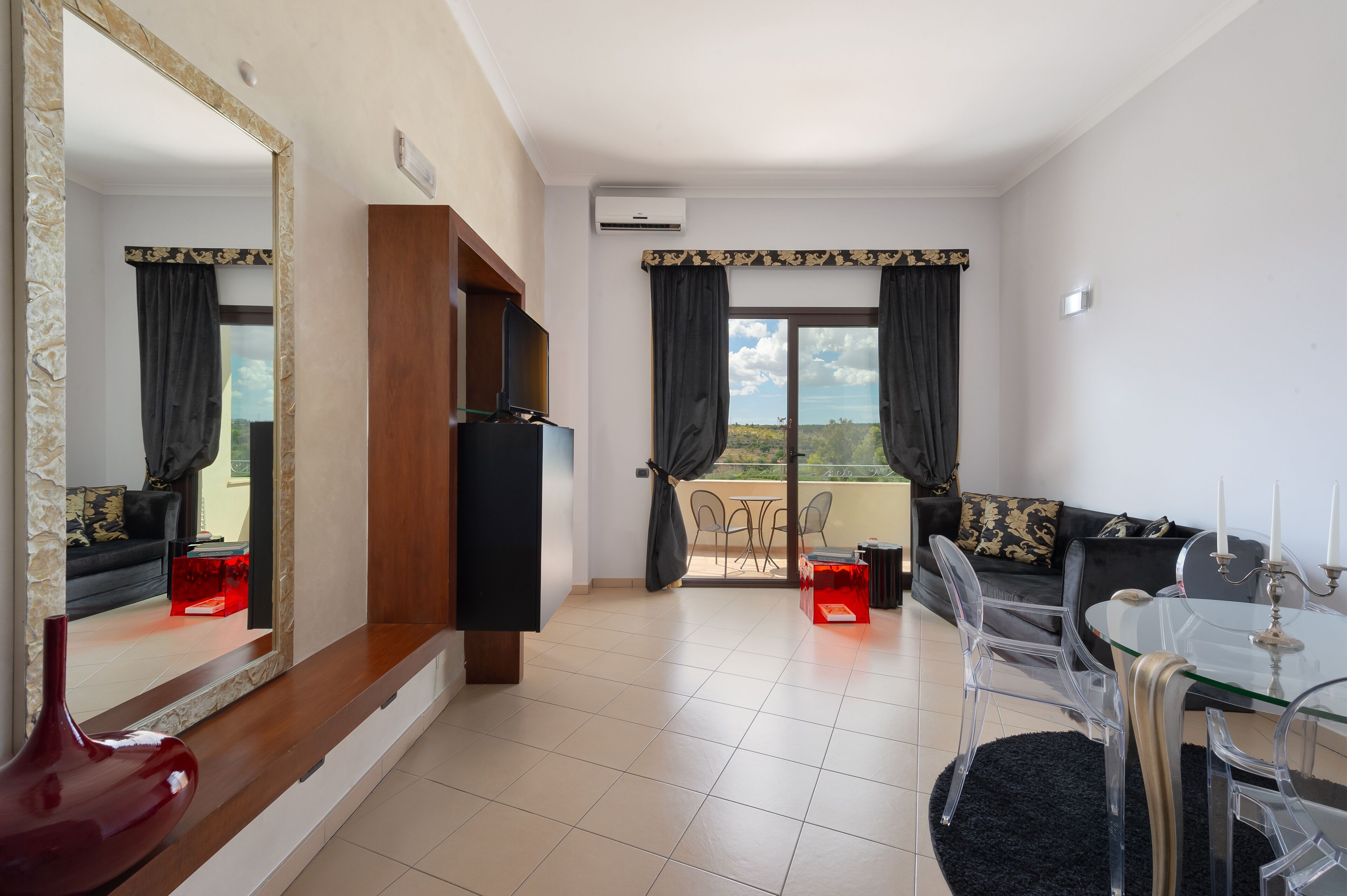 A modern living room with a glass dining table, clear chairs, a sofa, a TV, and a balcony with a view.
