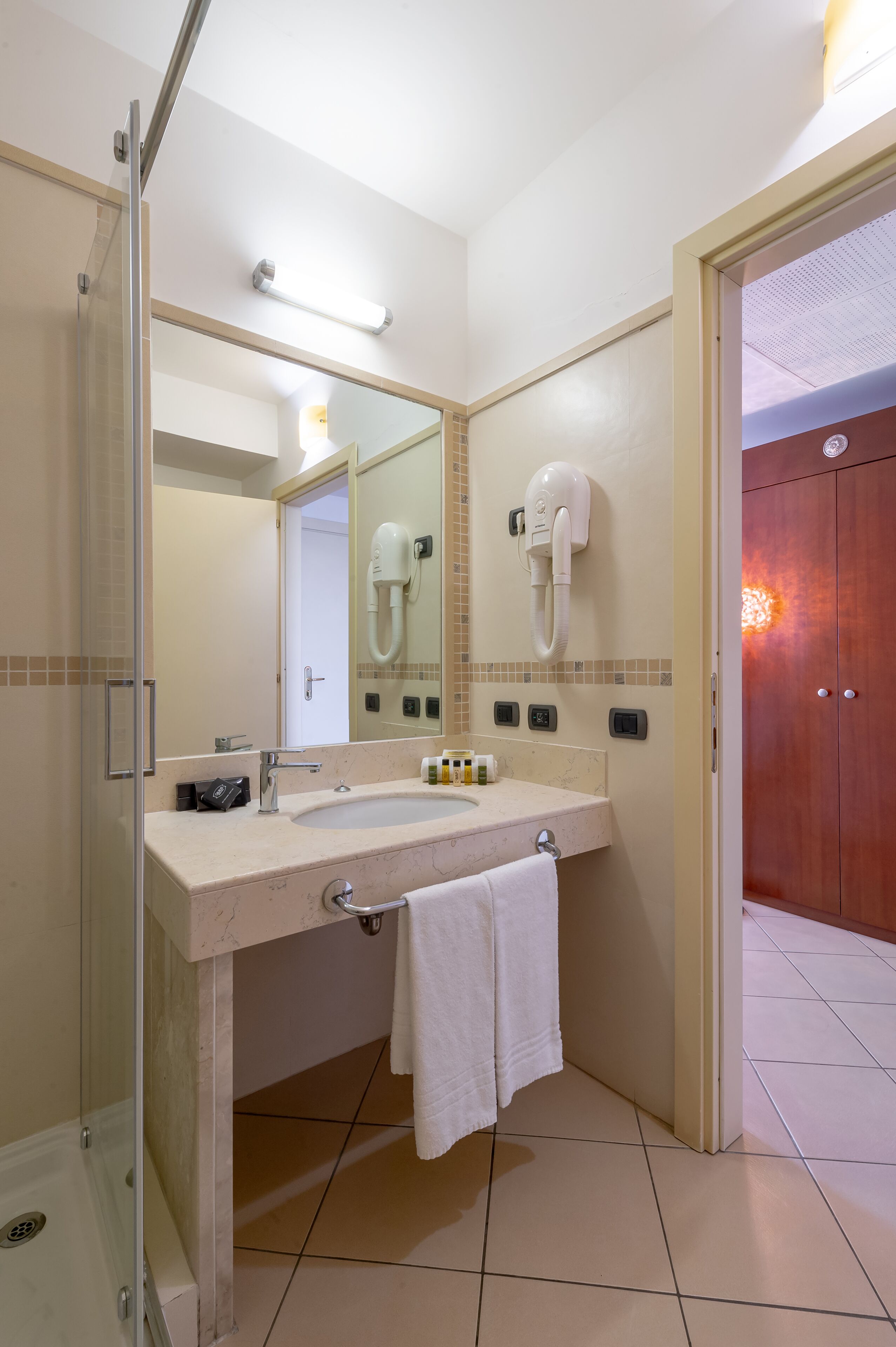 Ein Badezimmer mit Duschkabine aus Glas, Waschbecken mit Handtuch und einem an der Wand befestigten Fön.