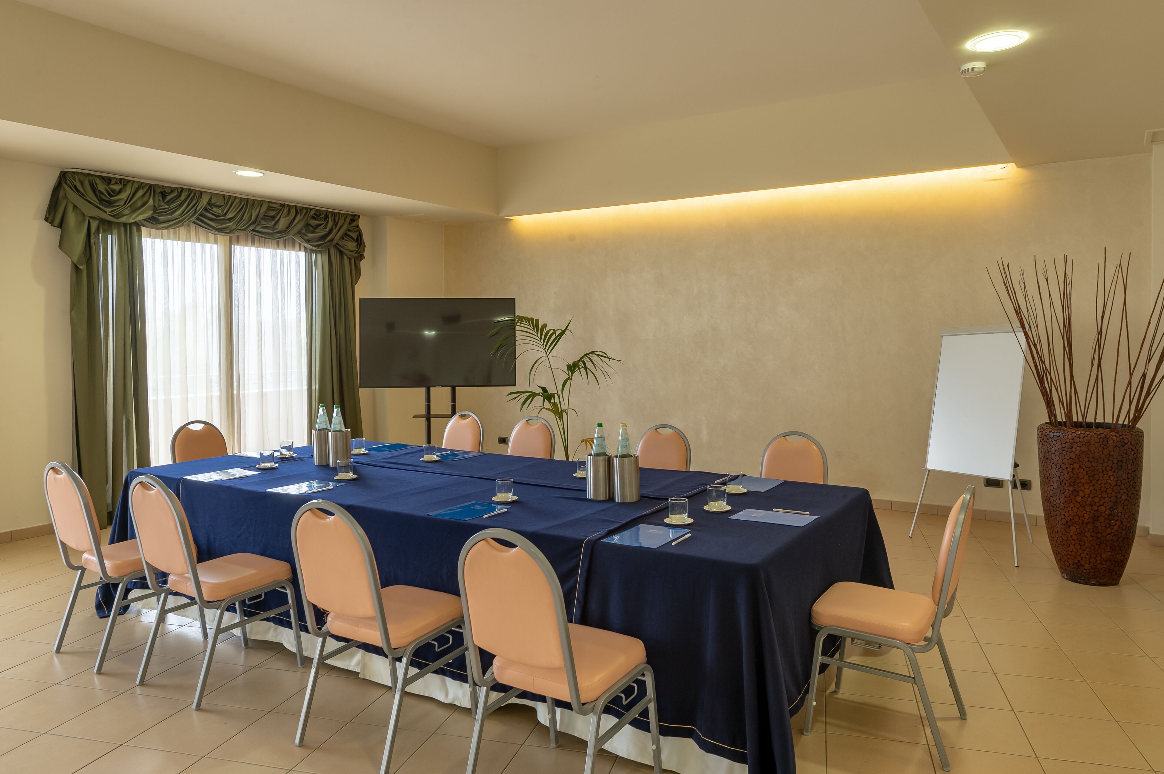 A conference room with a long table, chairs, a TV, a whiteboard, and a large potted plant.