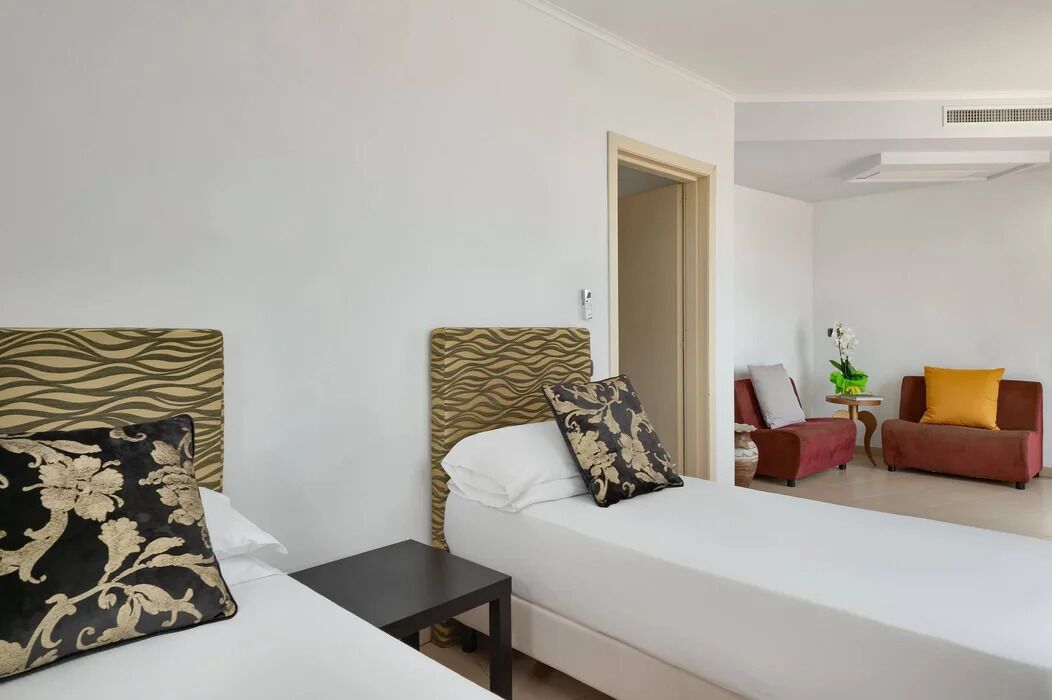 Habitación de hotel con dos camas, una mesita pequeña y una zona de estar con sofá y sillones.