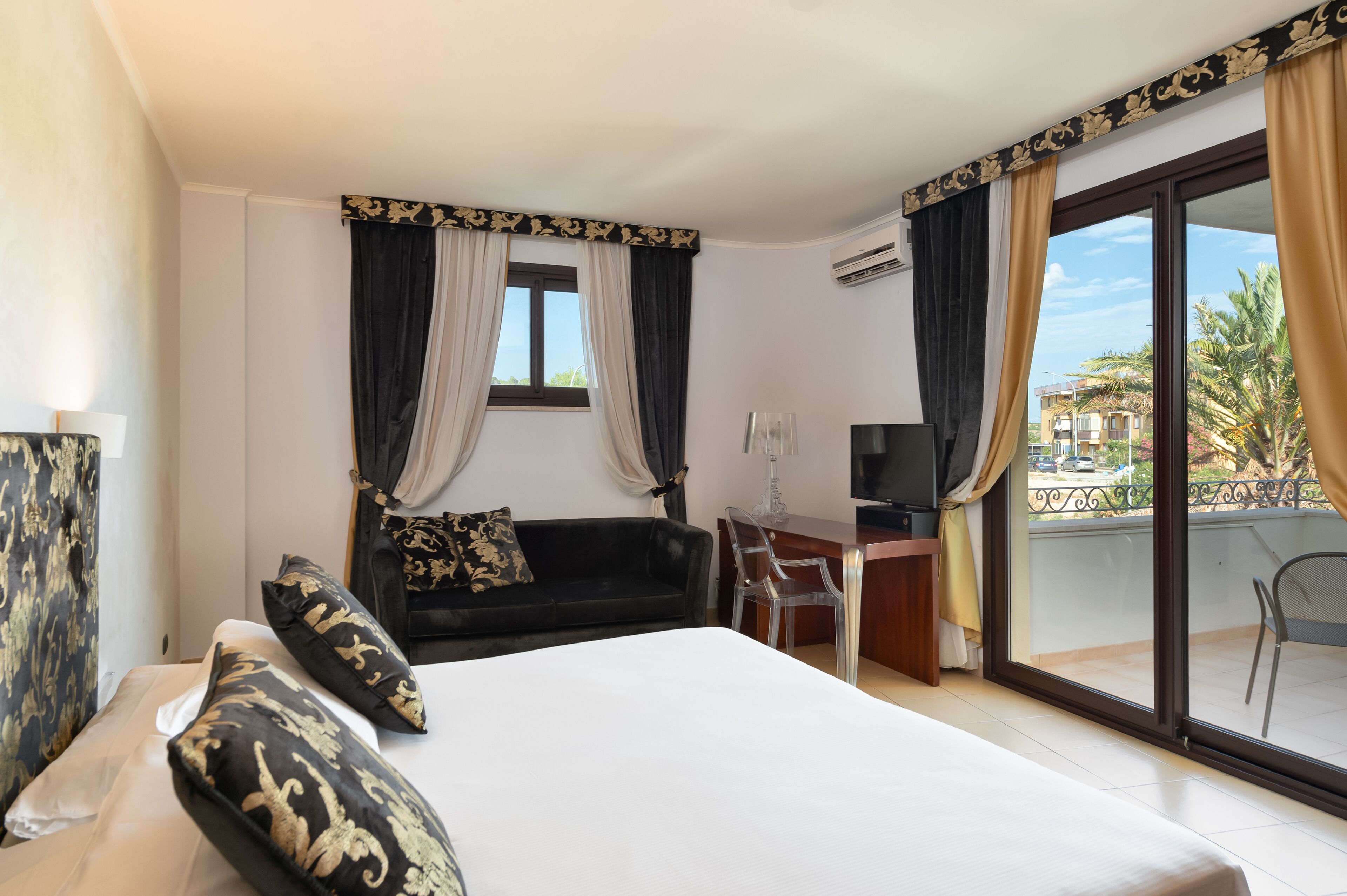 Habitación de hotel con cama, escritorio, televisor y balcón con vistas a palmeras.