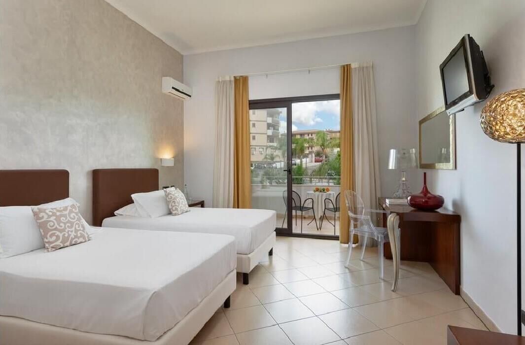 Habitación de hotel con dos camas, televisor, escritorio con lámpara y balcón con vistas.