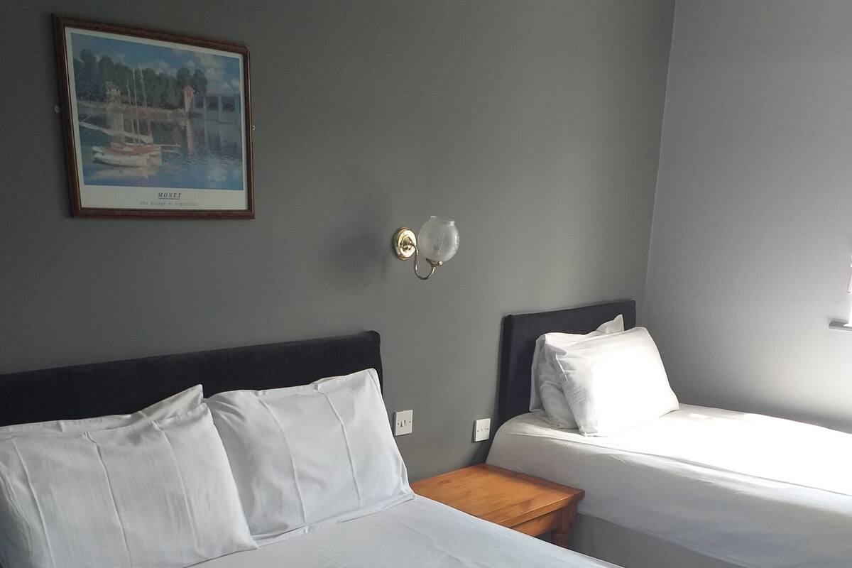 Ein Hotelzimmer mit zwei Einzelbetten, einem Nachttisch, einer Wandlampe und einem gerahmten Bild einer Brücke.
