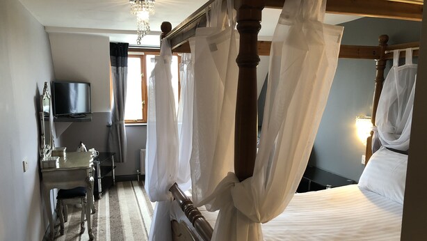 A bedroom with a four-poster bed, a bedside table, a chair, a television, and a window with curtains. - The Airman Hotel (Shefford)