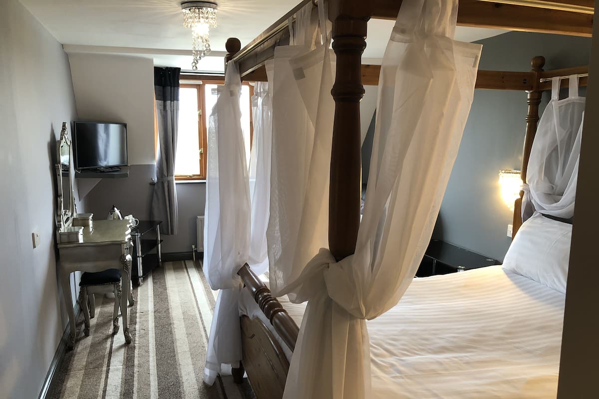 Ein Schlafzimmer mit einem Himmelbett, einem Nachttisch, einem Sessel, einem Fernseher und einem Fenster mit Vorhängen.