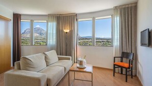 Habitación de hotel con sofá, una pequeña mesa con una botella y vasos, una silla y un televisor montado en la pared.