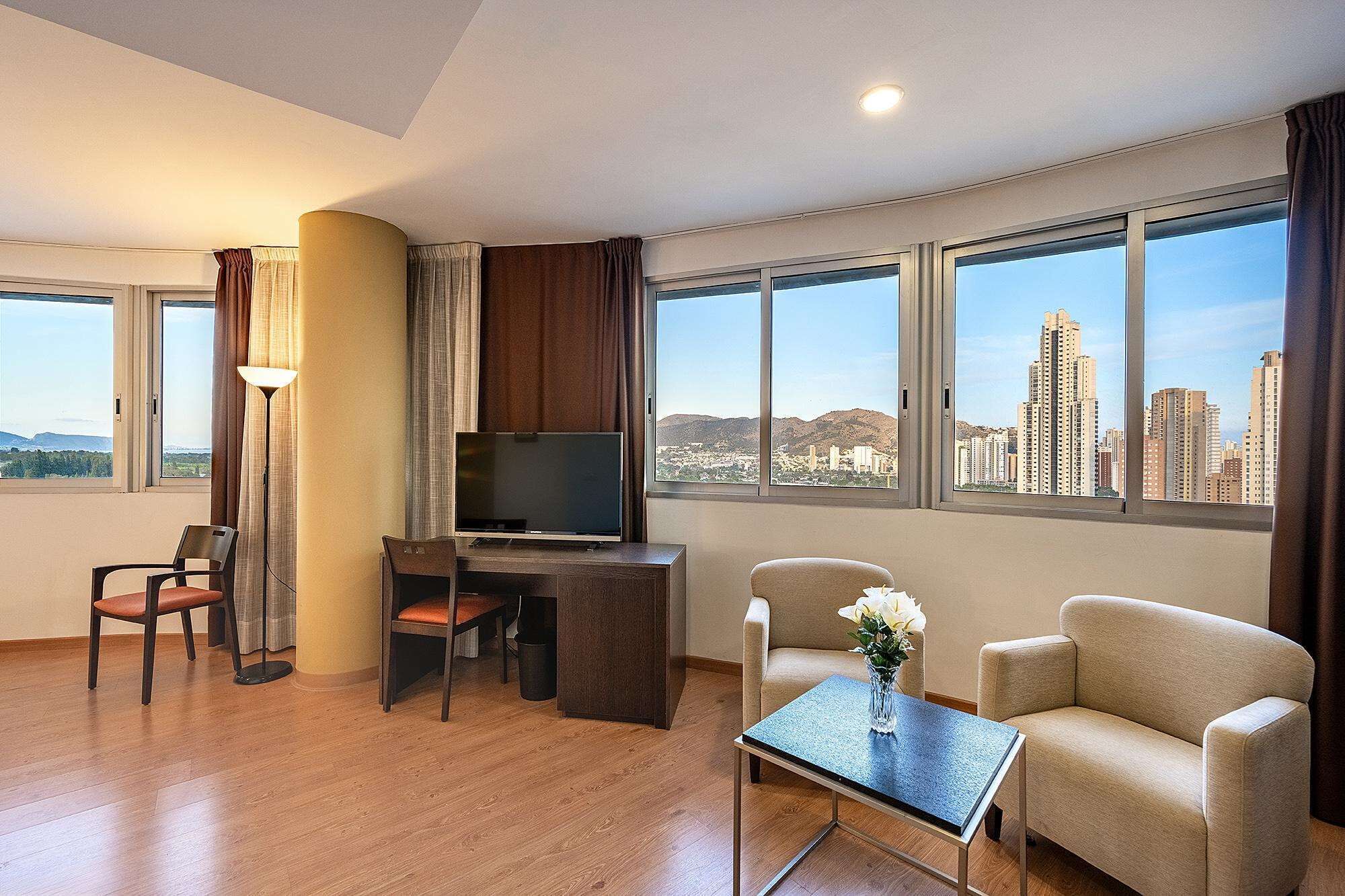 A hotel room with a desk, two chairs, a TV, a small table with a vase, and large windows offering a city view.