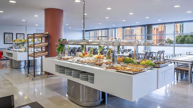 Montaje de bufé con diversos alimentos, un pilar grande y vistas de la ciudad a través de amplias ventanas.
