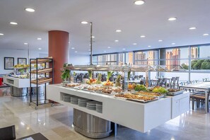 A buffet setup with various food items, a large pillar, and a view of the city through large windows. - Hotel La Estacion (Benidorm)