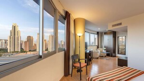 Una habitación de hotel moderna con un ventanal grande que ofrece vistas a la ciudad, un escritorio con silla, una cama con cubrecama a rayas y un armario.